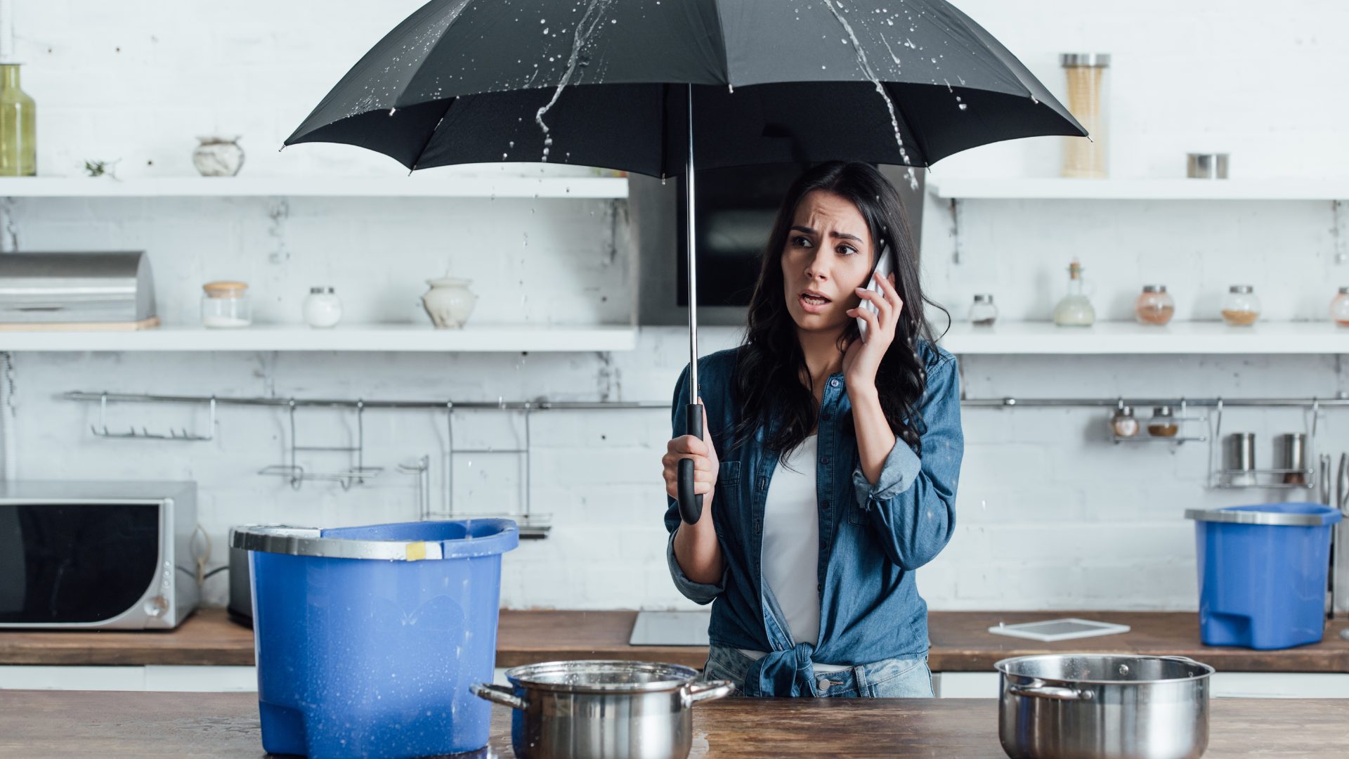 A woman in her kitchen, holding an umbrella as her roof leaks into a bucket and pots on the counter. She's on the phone calling for help.