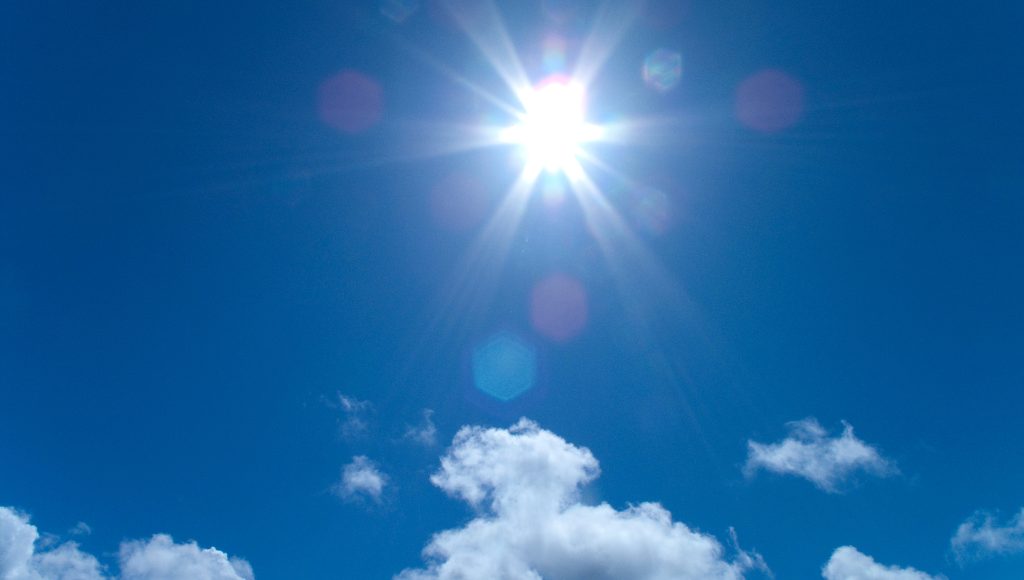 A bright sun shining in a blue sky surrounded by white fluffy clouds