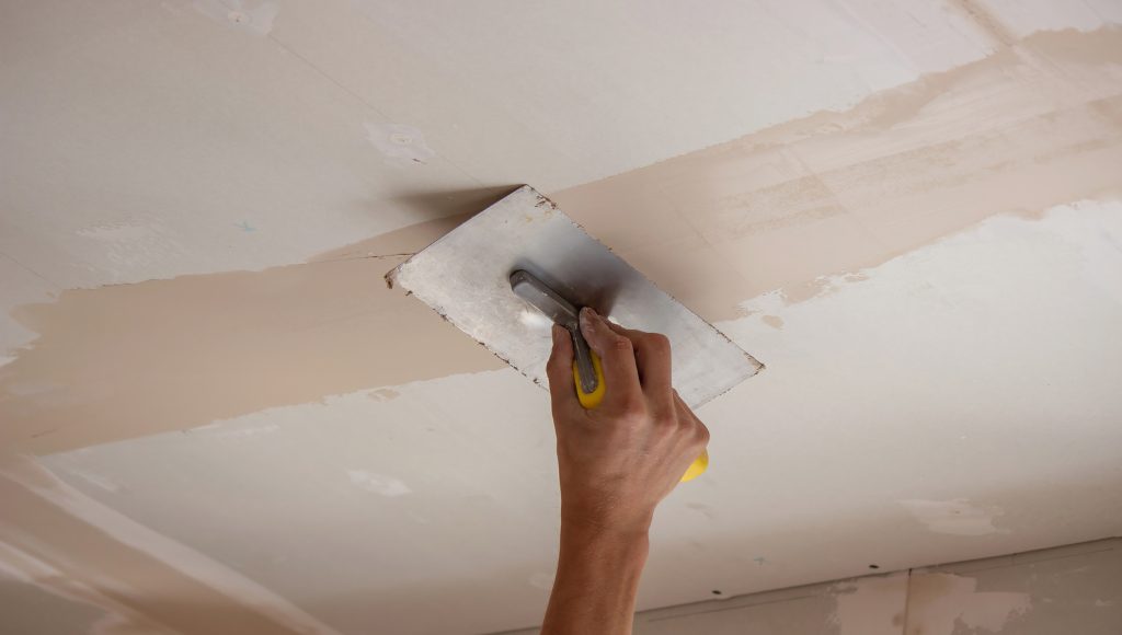 A man fixes a ceiling, smoothing a patch with a drywall trowel.