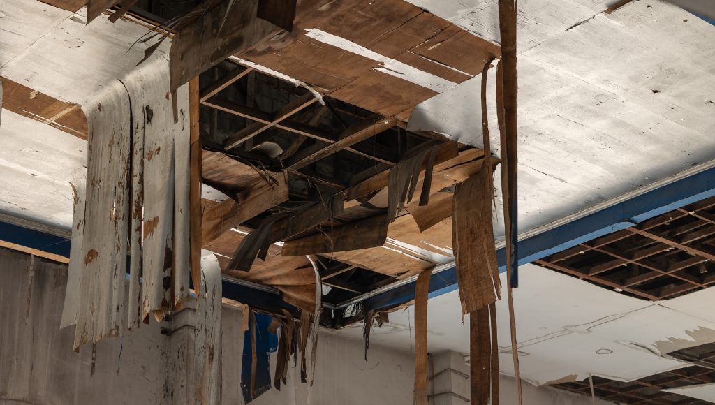 A severely weather and water damaged interior ceiling with sagging paint and wood, and missing ceiling pieces.