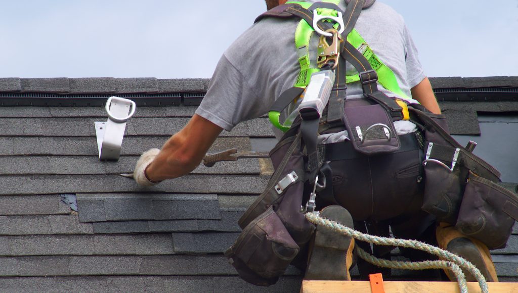 A roofer in a safety harness repairs damaged shingles.
