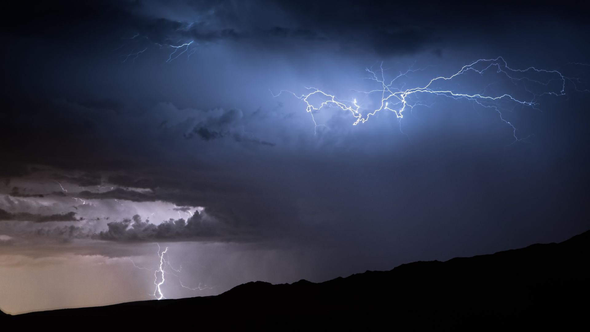 A night scene with lightning bolts flashing across a dark, cloudy sky above silhouetted hills.