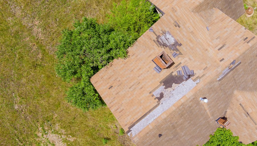 An aerial view of a roof covered in beige asphalt shingles that took damage during a recent storm with heavy rain and high winds. Shingles are missing in patches.