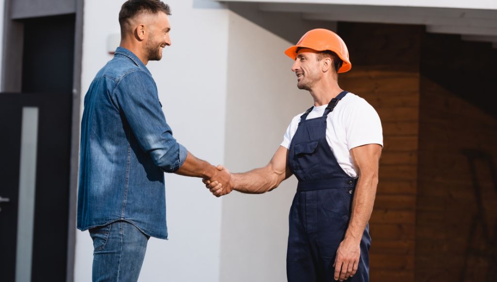 A homeowner in blue jeans and a blue denim shirt shakes the hand of a contractor in dark blue coveralls, white tshirt, and orange hard hat.