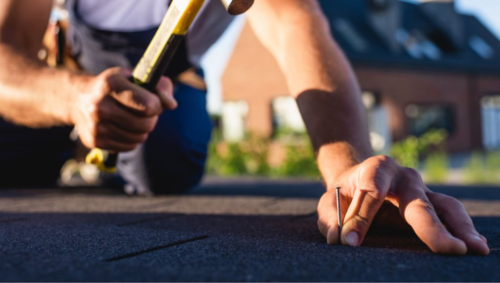 A roofer in blue coveralls and a white t-shirt repairs roof shingles with a hammer and nail.