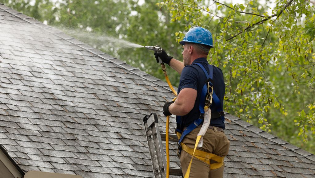 A certified Roof Maxx dealer in a blue hard hat and harness sprays the shingled roof of a residential home with Roof Maxx restoration treatment.