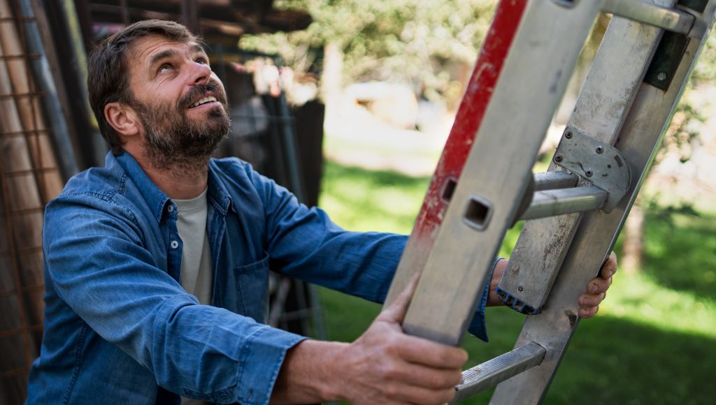 A homeowner in a blue denim overshirt and grey t-shirt places a ladder against the roof of his house.